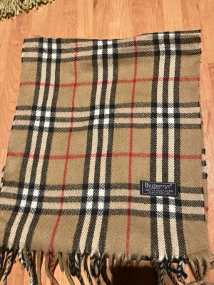 Beige rutig halsduk från Burberrys - Klassisk beige halsduk från Burberrys i 100% lammull med ikoniskt rutmönster i svart, vitt och rött. Halsduken har fransar i ändarna och en vävd Burberrys-etikett. Perfekt accessoar för att lyfta din outfit. Säljer den för 500 kr men är prutbar 