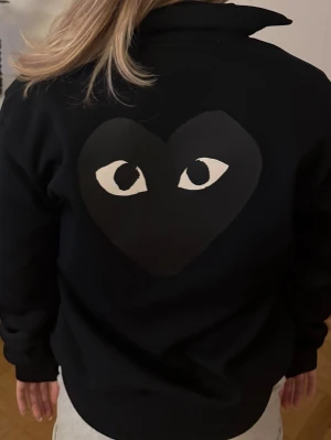 Svart zip hoodie med hjärtmotiv - Svart hoodie med dragkedja och högt krage. På ryggen finns ett stort svart hjärta med ögon, och samma motiv i mindre format på bröstet. Tillverkad i mjukt material, perfekt för en chill och stilren look.