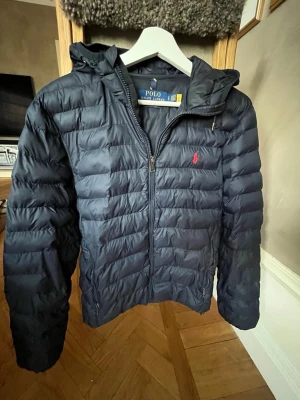 Mörkblå pufferjacka från Polo Ralph Lauren - Snygg mörkblå pufferjacka från Polo Ralph Lauren med huva och dragkedja framtill. Jackan har det klassiska röda Polo-logotrycket på bröstet och två sidofickor med dragkedja. Perfekt för kalla dagar och riktigt skön att bära. 