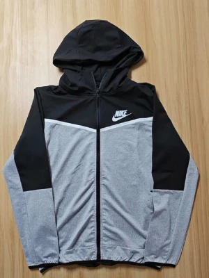 Nike svart och grått set med huva - Säljer ett snyggt tvådelat set från Nike med svart och grå zip-hoodie och matchande byxor. Jackan har huva, dragkedja och vit Nike-logga på bröstet. Byxorna har resår i midjan, snörning och logga på benet. Perfekt för en sportig och avslappnad stil.