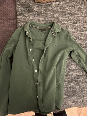 Grön skjorta från Tommy Hilfiger - Mörkgrön skjorta från Tommy Hilfiger med klassisk krage och vita knappar. Liten broderad logga på bröstet och diskret röd-vit detalj på insidan av kragen. Skjortan är i mjukt bomullsmaterial och har lång ärm.