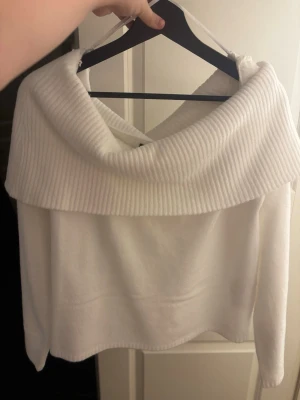Vit stickad tröja med bred krage - Säljer en vit stickad tröja med en bred, ribbad krage som kan bäras off-shoulder. Tröjan har en mjuk och mysig känsla, lång ärm och en något kortare passform. Perfekt för dig som gillar stilrena och trendiga plagg.