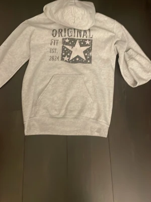 Grå hoodie från SCNDI - Snygg grå hoodie med huva och magficka. Tryck på ryggen med texten 'ORIGINAL FIT EST. 2024' och en stor stjärna omgiven av mindre stjärnor. Perfekt för en avslappnad och trendig stil.