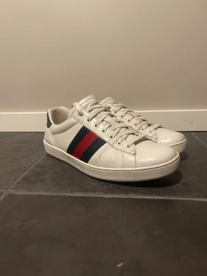 Gucci vita sneakers med ränder - Säljer ett par vita sneakers från Gucci med klassiska blå och röda ränder på sidan. Skorna har vita snören och detaljer i mörkblått ormskinnsmönster på hälen där Gucci-loggan är präglad. Tillverkade i läder och har en stilren, låg profil.