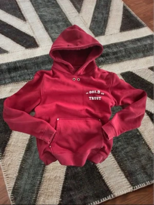 Röd hoodie från In Gold We Trust - Snygg röd hoodie från In Gold We Trust med vit text på bröstet och stor ficka framtill. Klassisk huva med dragsko och ribbade muddar vid ärmar och midja. Perfekt för en avslappnad streetwear-look.