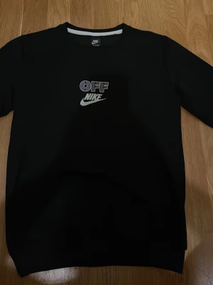 Svart sweatshirt från Nike - Svart sweatshirt från Nike med trycket 'OFF NIKE' i vitt och lila på bröstet. Klassisk rund halsringning och långa ärmar. Perfekt för en avslappnad streetwear-look.