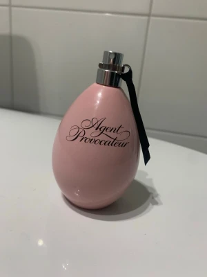 Agent Provocateur parfym rosa flaska - Agent Provocateur parfym i en elegant, äggformad rosa flaska med svart text och silverfärgad spraytopp. Flaskan har en svart banddetalj vid halsen som ger en lyxig känsla. Perfekt för dig som gillar snygg design på parfymhyllan.