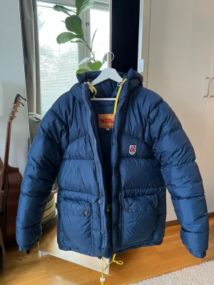 Blå pufferjacka från Fjällräven XS - Snygg blå pufferjacka från Fjällräven med huva och gula dragskor. Oversized 