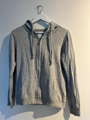 Ljusgrå zip hoodie med huva - En enkel och stilren ljusgrå hoodie med dragkedja framtill och två fickor. Hoodien har justerbara snören i huvan och är tillverkad i ett mjukt, tunt bomullsmaterial. Storlek: S