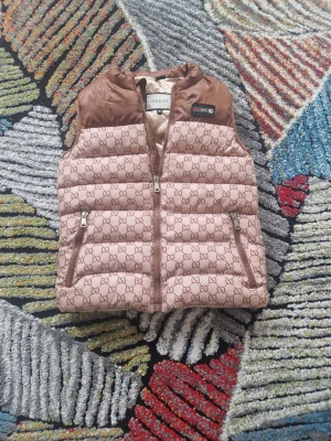 Beige dunväst från Gucci - Snygg beige dunväst från Gucci med klassiskt GG-monogram över hela västen och bruna detaljer upptill. Västen har dragkedja framtill, två fickor med dragkedja och en ikonisk Gucci-patch på bröstet. Perfekt för dig som vill ha en exklusiv och trendig look.