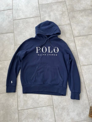 Marinblå hoodie från Polo Ralph Lauren - Snygg marinblå hoodie från Polo Ralph Lauren med stor vit logga framtill. Klassisk huva med dragsko och mjukt material som känns skönt mot huden. Perfekt för dig som gillar stilrena och tidlösa plagg med tydlig branding.