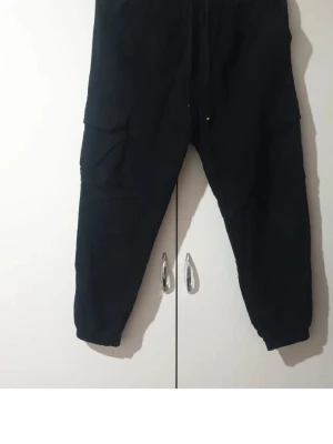 Svarta cargobyxor från Less Cost - Svarta cargobyxor från Less Cost med avslappnad passform och stora sidofickor. Tillverkade i en mjuk mix av 70% bomull, 25% polyester och 5% spandex för extra komfort och stretch. Perfekta för en casual och trendig look.