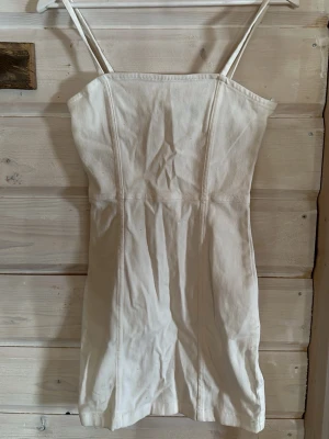 Vit kort klänning med smala band - Säljer en vit kort klänning med smala axelband och figurnära passform. Klänningen har en rak halsringning och dragkedja i ryggen för en snygg detalj. Materialet känns som bomull och ger en enkel, fräsch look.