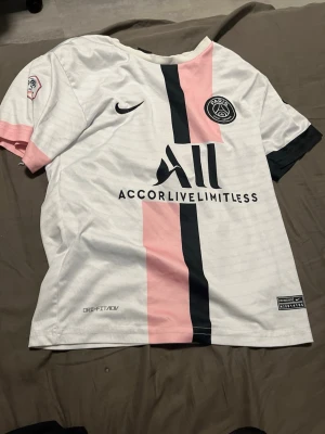 PSG fotbollströja Messi #30 Nike - Säljer en vit PSG matchtröja från Nike med rosa och svart rand på framsidan. Tröjan har PSG-logga, sponsortryck och Messi #30 på ryggen. Tillverkad i lätt och ventilerande material, perfekt för fotboll eller träning.