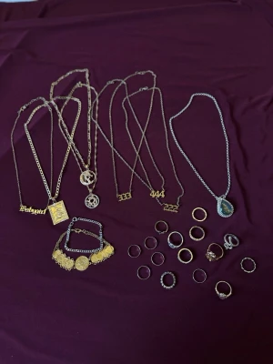 Mix av halsband, armband och ringar i guld och silver - Samling med flera halsband, armband och ringar i både guld- och silverfärg. Halsbanden har olika hängen som siffror, text, medaljonger och stenar. Ringarna varierar i design, vissa med stenar, ormmotiv och blommor. Perfekt för dig som gillar att mixa och matcha accessoarer.