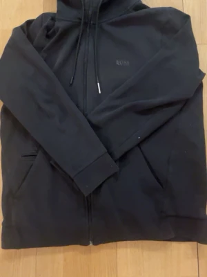 Svart hoodie med dragkedja från BOSS - Säljer en svart hoodie från BOSS med dragkedja framtill och två fickor. Klassisk design med huva och justerbara snören. Diskret BOSS-logga på bröstet. Tillverkad i mjukt material som känns skönt mot huden. Perfekt för en avslappnad och stilren look.