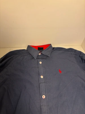 Blårutig skjorta från polo - Klassisk blårutig skjorta från polo med röd broderad logga på bröstet. Skjortan har vit-röda knappar och en snygg kontrast med röd insida vid kragen. Perfekt för dig som gillar stilrena detaljer och tidlös design.