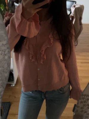 Ljusrosa kofta med volangdetaljer - Supersöt ljusrosa kofta med knappar framtill och volangdetaljer längs knappraden och axlarna. Långärmad modell med ribbade muddar och nederkant. Perfekt för dig som gillar en feminin och trendig stil.