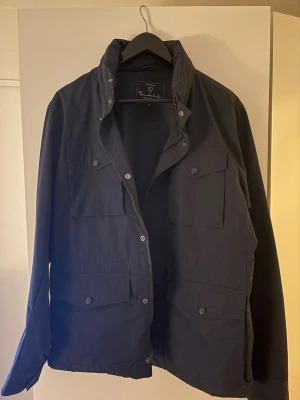 Mörkblå fieldjacket från Bondelid - Snygg mörkblå fieldjacket från Bondelid med fyra stora fickor framtill och hög krage. Jackan har dragkedja och tryckknappar samt en stilren, minimalistisk design. Tillverkad i ett lätt och vindtätt material som passar perfekt för blåsiga dagar.