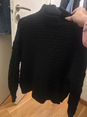 Svart ribbstickad tröja med polokrage - Säljer en svart ribbstickad tröja med hög polokrage och långa ärmar. Tröjan har en avslappnad passform och är gjord i ett mjukt, tjockt material som passar perfekt för kyliga dagar. Enkel och stilren design som är lätt att matcha.