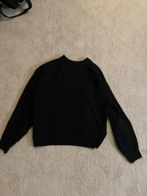 Svart stickad tröja med rund hals - En enkel och stilren svart stickad tröja med rund hals och långa ärmar. Tröjan har en rak passform och ribbade muddar vid ärmslut och nederkant. Perfekt att matcha med jeans eller kjol för en clean look.