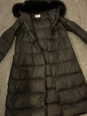 Svart lång dunjacka med pälshuva - Säljer en svart lång dunjacka från BR med stor huva och fluffig pälskant. Jackan har quiltad design, knappar och dragkedja framtill. Perfekt för kalla vinterdagar och riktigt varm tack vare dunfyllningen och den långa modellen.