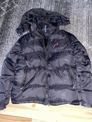 Svart pufferjacka från Polo Ralph Lauren - Snygg svart pufferjacka från Polo Ralph Lauren med huva och dragkedja framtill. Jackan har två stora fickor med dragkedja och den klassiska röda loggan broderad på bröstet. Perfekt för kalla vinterdagar och riktigt skön att bära.