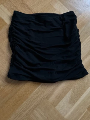 Svart tight kortkjol med rynkade sidor - Säljer en svart tight kortkjol med rynkade sidor. Kjolen har en enkel och stilren design som passar perfekt till många olika outfits. Materialet känns mjukt och stretchigt vilket gör den bekväm att bära.