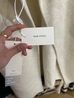 Beige lång kappa med fickor - Stilren beige kappa från New Style med rak passform och stora framfickor. Kappan har en minimalistisk design med knäppning framtill och rundad krage. Perfekt för dig som gillar en clean och modern look. Kappan är 70% ull