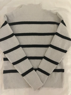 Grå stickad tröja med svarta ränder 🤍🖤 - Säljer en ribbstickad grå tröja från Vila med svarta horisontella ränder. Tröjan har hög hals och långa ärmar, perfekt för kyligare dagar. Materialet är mjukt och stretchigt, vilket gör den riktigt skön att bära.💞