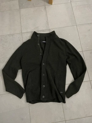 Mörkgrön kofta från Barbour - Snygg mörkgrön kofta från Barbour med både dragkedja och knappar framtill. Tröjan har ribbad krage, muddar och två fickor. Tillverkad i mjukt stickat material som känns riktigt skönt mot huden. Perfekt för lager på lager och stilren look.