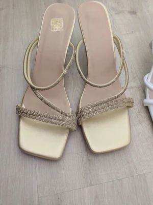 Guldiga pumps med strass - Säljer ett par eleganta guldiga pumps från med fyrkantig tå och smala remmar dekorerade med strass. Skorna har en låg klack på 6cm och öppen häl, perfekta för att lyfta din outfit med lite extra bling. Stl 36, men passar mig med stl 37. Råkat beställa dubbletter, det jag skickar kommer vara oöppnat och oanvänt💗