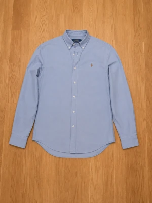 Blå skjorta från Polo Ralph Lauren - Klassisk ljusblå skjorta från Polo Ralph Lauren med button-down krage och broderad logga på bröstet. Skjortan har lång ärm, knappar framtill och är tillverkad i bomull. Perfekt för en clean och stilren look.