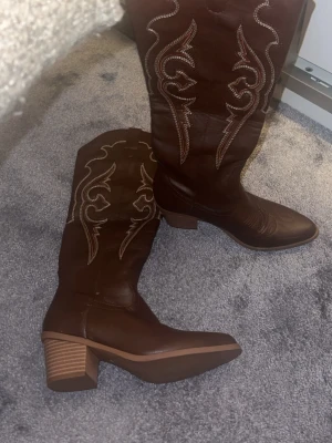 Bruna cowboy boots med broderi - Snygga bruna cowboy boots med klassiskt vitt broderi längs skaftet och spetsig tå. Bootsens klack är bred och stabil, och de har en autentisk westernstil. Perfekta för dig som vill sticka ut med en cool look.