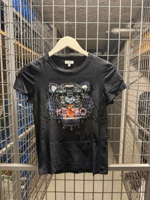 Svart Kenzo t-shirt med tigertryck - Cool svart t-shirt från Kenzo med ikoniskt tigertryck och färgglada detaljer framtill. Klassisk rund hals och korta ärmar. Perfekt för dig som vill sticka ut med en statement-tee från ett välkänt märke.