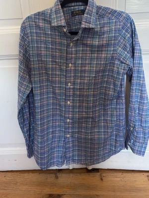 Rutig skjorta från Polo Ralph Lauren - Klassisk rutig skjorta från Polo Ralph Lauren i blå, grön och vit färgskala. Skjortan har lång ärm, knappar framtill och en avslappnad passform. Perfekt till jeans eller chinos för en stilren look.