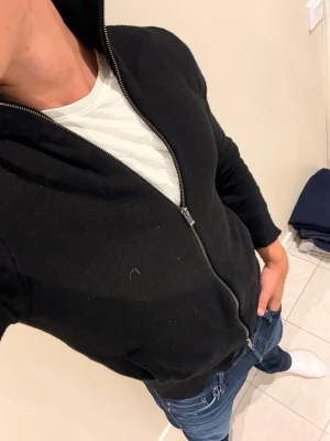 Svart zip-hoodie med huva - En svart hoodie med dragkedja och huva. Klassisk modell med långa ärmar och fickor framtill. Perfekt att slänga på sig över en t-shirt. Materialet ser ut att vara bomull eller bomullsblandning. Enkel och stilren design utan synligt märke.