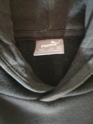 Svart hoodie från Puma med logga - Svart hoodie från Puma med stor vit logga på bröstet. Klassisk modell med huva och magficka. Mjuk insida och ribbade muddar vid ärmslut och nederkant. Perfekt för en avslappnad och sportig stil.
