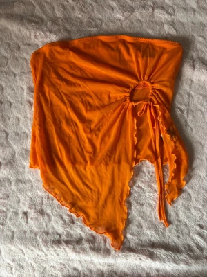 Orange asymmetrisk tubtopp - Trendig orange tubtopp med asymmetrisk nederkant och snygg dragsko-detalj framtill. Toppen är ärmlös och har en cool, edgy look som passar perfekt till sommaren. Materialet känns lätt och luftigt.