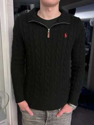 Svart kabelstickad tröja Polo Ralph Lauren - Svart kabelstickad tröja från Polo Ralph Lauren med halv dragkedja och röd broderad logga på bröstet. Tröjan har ribbade muddar och hög krage, perfekt för lager-på-lager. Klassisk och stilren design som funkar till det mesta. Den är lite noppig 