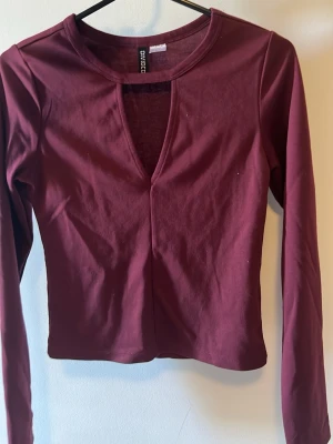 Vinröd långärmad topp från H&M Divided - Vinröd långärmad topp från H&M Divided med djup v-ringning och figurnära passform. Tillverkad i mjukt och stretchigt material, perfekt att styla med jeans eller kjol. Enkel men snygg design som passar till många olika looks.
