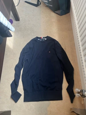 Mörkblå sweatshirt från Tommy Hilfiger - Mörkblå sweatshirt med rund hals från Tommy Hilfiger. Tröjan har en liten broderad logotyp på bröstet och ribbade muddar vid ärmslut och nederkant. Enkel och stilren design som passar till det mesta. Priset kan diskuteras 