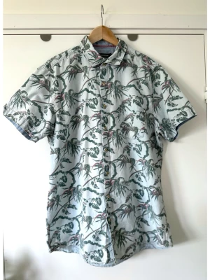 Dressmann - blå mönstrad kortärmad skjorta - XL - Snygg blå kortärmad skjorta från Dressman i storlek XL med blommigt palm-mönster i grönt och brunt på ljusblå bakgrund. Klassisk krage och knäppning framtill. Perfekt för en avslappnad och trendig look. Finvävd jeans / denim- tyg i 100% bomull, så bra material som andas. Perfekt inför vår och sommar! Mycket gott skick!