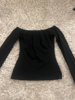 Svart offshoulder topp - Snygg svart offshoulder topp med lång ärm. Perfekt för dig som gillar en stilren och enkel look. Toppens passform är tight och den har en klassisk, rak skärning. Enkel att matcha med både jeans och kjol.