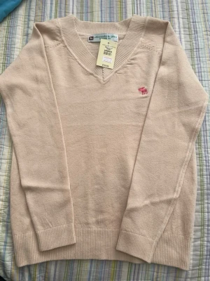 Beige cashmere tröja Abercrombie - Beige stickad tröja från Abercrombie & Fitch med V-ringning och ribbade muddar. Tröjan har en broderad rosa älg-logga på bröstet och är långärmad. Perfekt för lager-på-lager och enkel att matcha med jeans eller kjol. 100% cashmere
