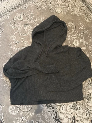 Mörkgrå croppad hoodie - Mysig mörkgrå hoodie med croppad passform och ribbad struktur. Tröjan har huva med dragsko och långa ärmar. Perfekt för en avslappnad och trendig stil.