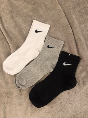 Nike strumpor i tre färger - Tre par klassiska Nike strumpor i vitt, grått och svart med broderad logga på skaftet. Strumporna är ribbade och tillverkade i mjuk bomullsblandning som sitter skönt på foten. Perfekta för en sportig och avslappnad stil. Strl 36-42 alla 3 för 100kr