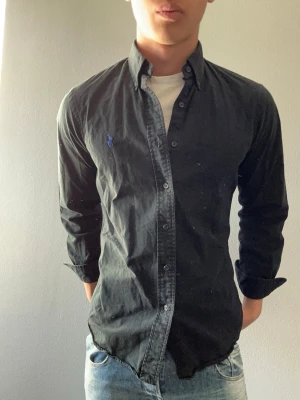 Svart skjorta från Ralph Lauren - Snygg svart skjorta från Ralph Lauren med klassisk krage och blå broderad logga på bröstet. Skjortan har lång ärm, knäppning framtill och är tillverkad i bomull. Perfekt för en stilren look.