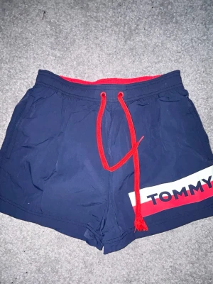 Marinblå badbyxor från Tommy Hilfiger - Snygga marinblå badbyxor från Tommy Hilfiger med röd dragsko och logga framtill. Bakficka med färgblock i rött och vitt samt liten logotyp. Klassisk modell i snabbtorkande material, perfekt för stranden eller poolen. Det finns tecken på användning, rivning på baksidan. Ca 32 cm långa