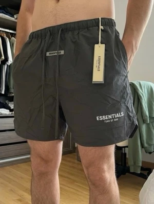 Svarta shorts från Essentials Fear of God - Svarta shorts från Essentials Fear of God med vit logga på benet. De har elastisk midja med snörning och är tillverkade i ett lätt och bekvämt material, perfekta för sommaren. Snygg och enkel design som passar till många olika stilar.